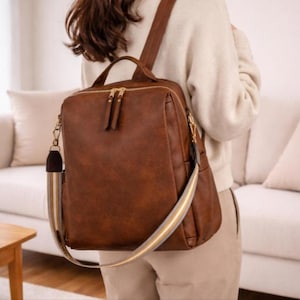 Puede incluir: Una mochila de cuero marrón con asa superior y cremalleras doradas. La correa ajustable para el hombro está a rayas beige, blanco y dorado. La bolsa se lleva sobre el hombro.