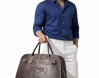 May include: A brown leather travel bag with a zip pocket and two handles. The bag has a rectangular shape with rounded edges. The bag is held by a person wearing a blue shirt and white trousers.