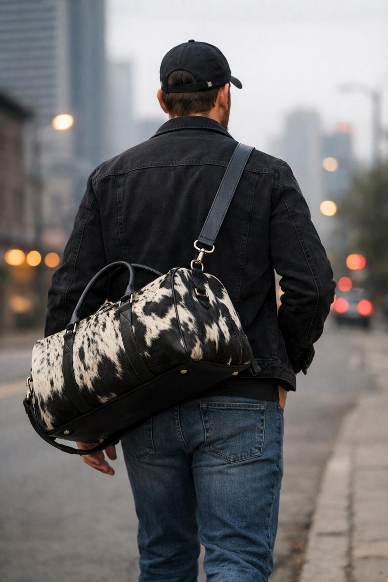 May include: A duffel bag featuring a black and white cowhide pattern with black leather trim, black handles, and a detachable black shoulder strap. The bag has a structured base with metal feet and silver-toned hardware attachments for the strap.