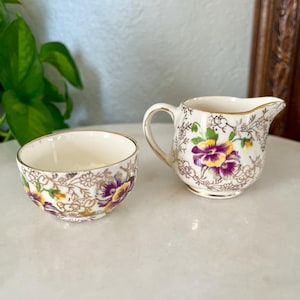May include: A vintage cream and sugar set. The small creamer and bowl are white with a gold rim and decorated with purple and yellow floral patterns. The set is likely porcelain.