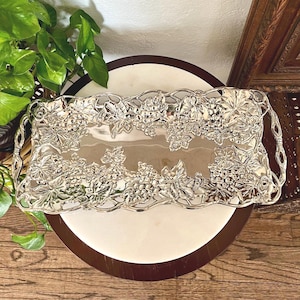 May include: A silver-colored rectangular serving tray with a detailed grape and leaf design around the edges. The tray has handles on each end and is placed on a white and brown surface.