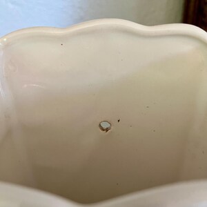 May include: Cream-colored ceramic planter with a scalloped rim. The interior has a small drainage hole. The planter's surface has minor imperfections, adding to its vintage charm. A decorative piece for plants.