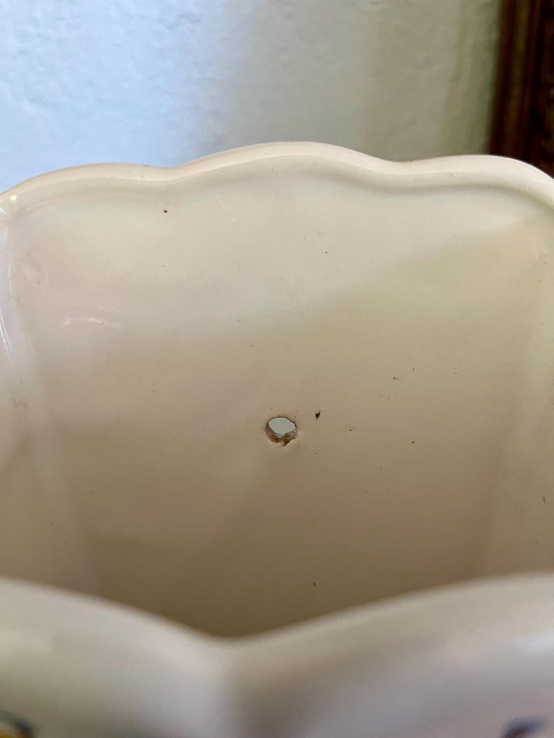 May include: Cream-colored ceramic planter with a scalloped rim. The interior has a small drainage hole. The planter's surface has minor imperfections, adding to its vintage charm. A decorative piece for plants.