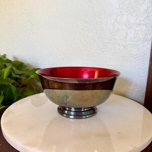 May include: A small, round bowl featuring a vibrant, glossy red interior and a metallic exterior with a weathered, greenish-brown patina. The bowl is supported by a circular pedestal base, creating a distinct contrast between the smooth, bright red inner surface and the textured, aged appearance of the outer metal body.