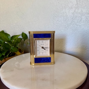 May include: A rectangular Bulova quartz clock featuring a polished gold-tone metal frame with vertical fluted side panels. The clock face is white with black Roman numerals and hands, set against a deep blue marble-patterned inlay background.