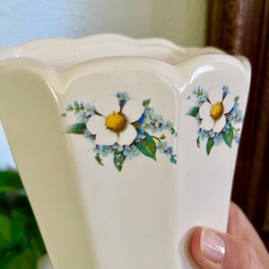 May include: Cream-colored, hexagonal vase with a scalloped rim. The vase is decorated with white flowers with yellow centers and small blue flowers with green leaves. The vase is held by a person.
