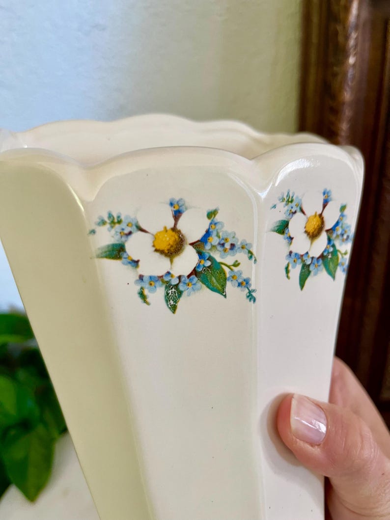 May include: Cream-colored, hexagonal vase with a scalloped rim. The vase is decorated with white flowers with yellow centers and small blue flowers with green leaves. The vase is held by a person.