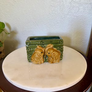 May include: A rectangular ceramic planter featuring a dark green bamboo-textured exterior. Two light brown elephant figures are molded onto the front, facing each other. The interior of the planter is a deep green, and the overall design has a glossy finish.