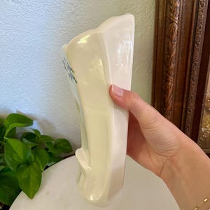 May include: A vintage, off-white ceramic vase with a flared top and a curved, sculpted base. Delicate blue floral accents adorn the front. The vase is held by a hand, showcasing its smooth, glossy finish. A classic piece for home decor.