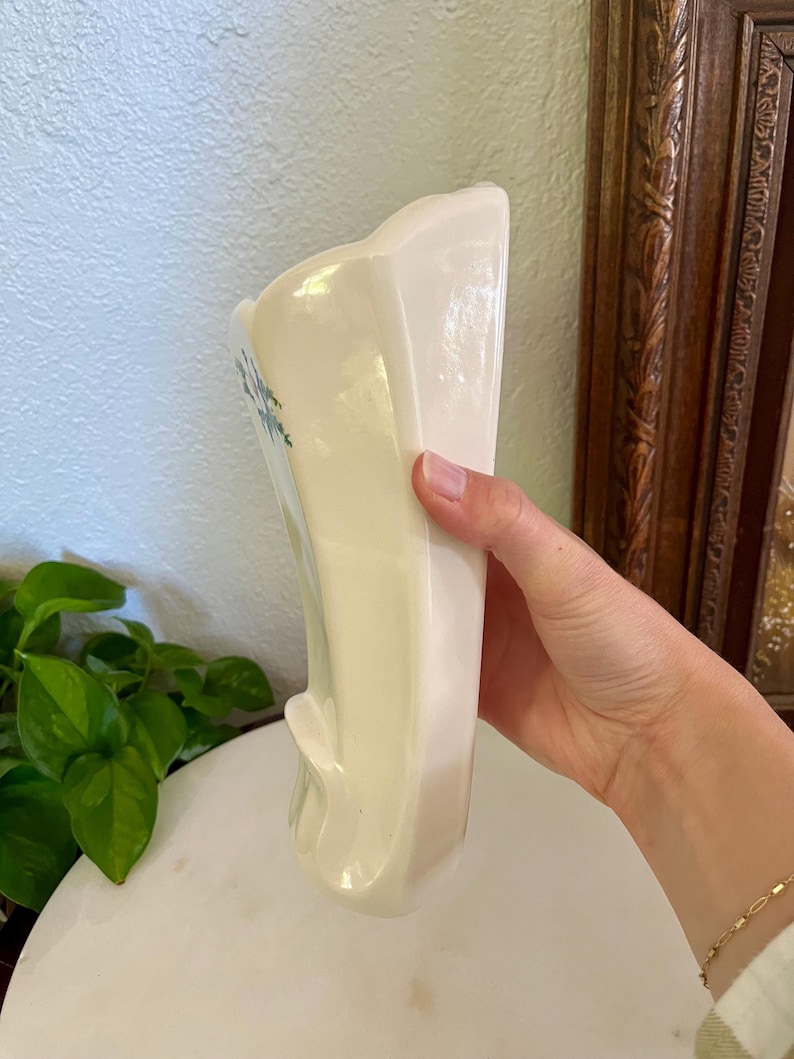 May include: A vintage, off-white ceramic vase with a flared top and a curved, sculpted base. Delicate blue floral accents adorn the front. The vase is held by a hand, showcasing its smooth, glossy finish. A classic piece for home decor.