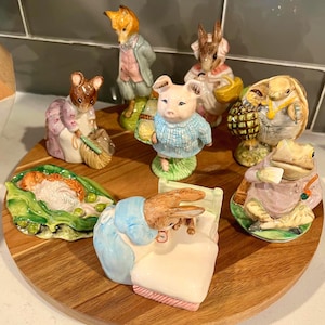 May include: A collection of ceramic figurines featuring anthropomorphic animals. The figurines include a fox, rabbit, pig, mouse, and frog, each dressed in detailed clothing and posed in various scenes. The figurines are arranged on a wooden serving tray.