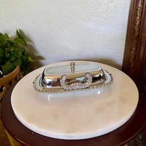 May include: A rectangular silver-toned butter dish with a reflective lid and a matching base. The lid features a small central knob, while the base has an ornate, scalloped edge with intricate embossed detailing along the rim and handle.