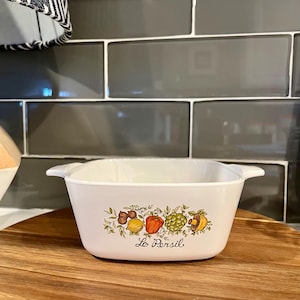 May include: White rectangular casserole dish with a colorful fruit and vegetable design. The dish has two handles and the words "Le Persil" are written on the front. The dish is sitting on a wooden cutting board.