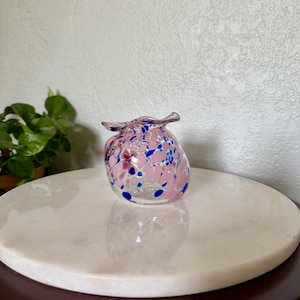 May include: A small, rounded glass vase with a ruffled, flared rim. The clear glass body is decorated with a speckled pattern of opaque light pink and vibrant blue spots, creating a colorful, multi-layered appearance throughout the vessel.