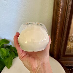 May include: A white ceramic creamer with a floral design. The creamer is held in a hand, showcasing its glossy finish and delicate details. The creamer has a small handle and a spout for pouring.