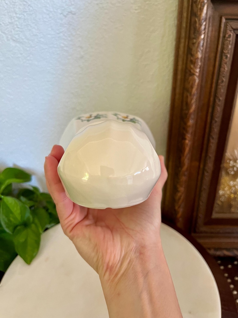 May include: A white ceramic creamer with a floral design. The creamer is held in a hand, showcasing its glossy finish and delicate details. The creamer has a small handle and a spout for pouring.