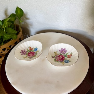 May include: Two white porcelain dishes shaped like scallop shells, each featuring a scalloped edge and a gold-trimmed rim. The center of each dish is decorated with a colorful floral arrangement including pink, purple, and blue flowers.