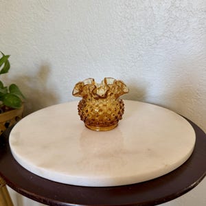 May include: A small amber-colored glass vase featuring a hobnail texture on the exterior and a ruffled, scalloped rim. The vessel has a rounded body and a translucent finish that catches the light, showcasing the raised dot pattern.