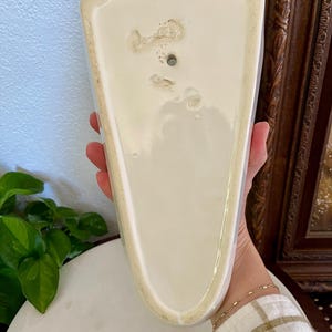 May include: A vintage, off-white ceramic wall pocket with a scalloped top edge. The pocket has a triangular shape and a hole for hanging. The back shows signs of age and wear, adding to its antique charm. A decorative piece for displaying small plants or flowers.