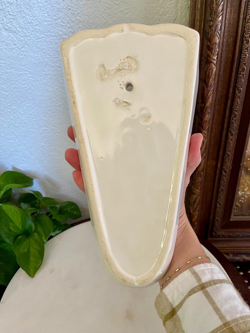 May include: A vintage, off-white ceramic wall pocket with a scalloped top edge. The pocket has a triangular shape and a hole for hanging. The back shows signs of age and wear, adding to its antique charm. A decorative piece for displaying small plants or flowers.