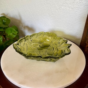 May include: A translucent olive green glass bowl featuring a detailed embossed pattern of overlapping leaves and small acorns. The bowl has a scalloped, irregular rim that follows the organic shape of the foliage design, creating a textured surface.