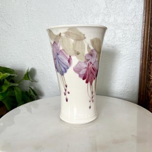 May include: A cream-colored ceramic vase with a floral design. The vase features purple, pink, and tan flowers. The vase is approximately 8 inches tall and has a flared top.