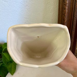 May include: Cream-colored ceramic vase with a unique, curved shape. The vase has a wide, open top and a smooth, glossy finish. The interior is a darker cream color. The vase is likely for decorative purposes.