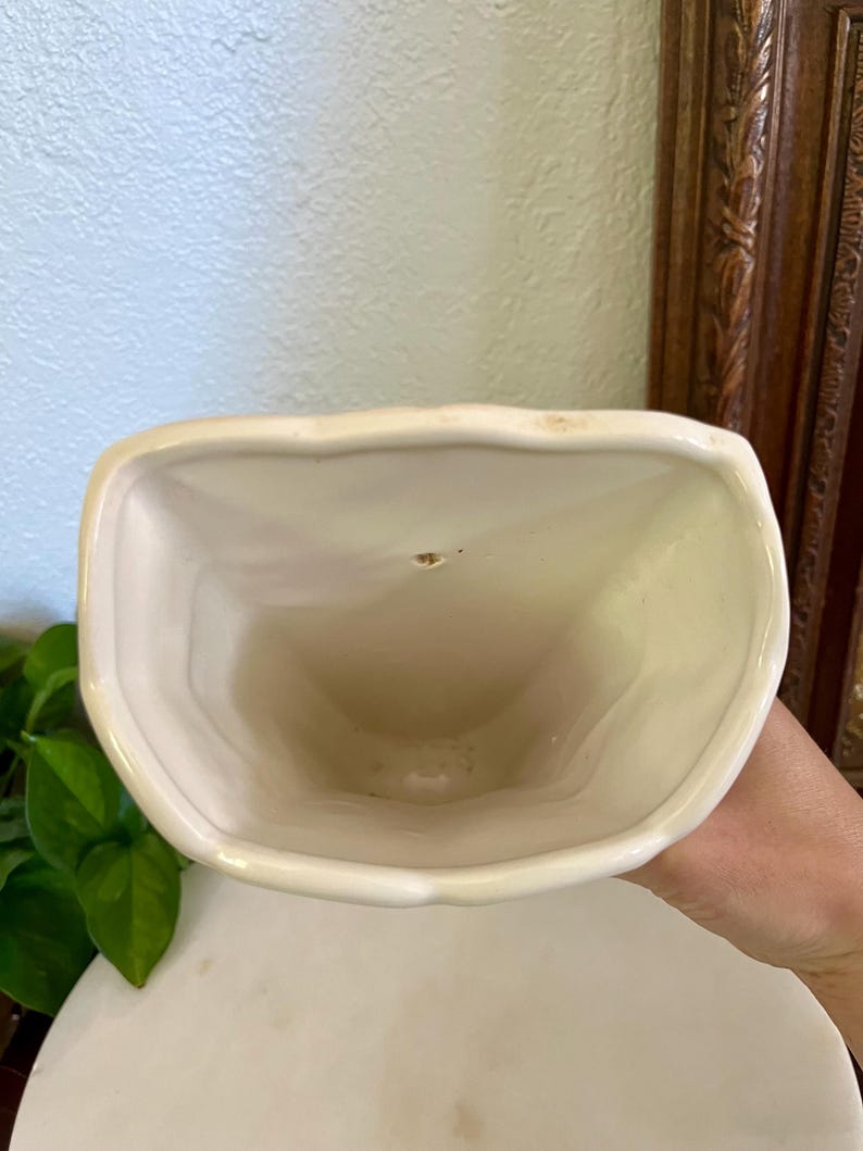 May include: Cream-colored ceramic vase with a unique, curved shape. The vase has a wide, open top and a smooth, glossy finish. The interior is a darker cream color. The vase is likely for decorative purposes.