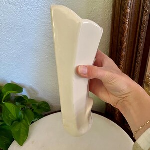 May include: A cream-colored, ceramic vase with a unique, asymmetrical shape. The vase has a smooth, glossy finish and a decorative, curved element on the side. The vase is held by a person.