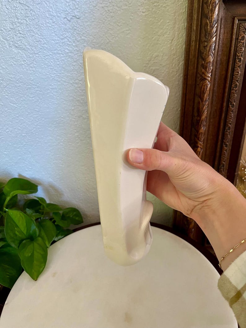 May include: A cream-colored, ceramic vase with a unique, asymmetrical shape. The vase has a smooth, glossy finish and a decorative, curved element on the side. The vase is held by a person.