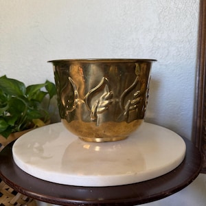 May include: A polished brass bowl featuring a repeating embossed leaf pattern around the exterior. The bowl has a wide, circular opening and a smooth, rounded base, showcasing a metallic gold finish with subtle variations in tone and texture.