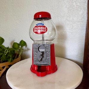 May include: A red gumball machine with a clear glass globe featuring the Dubble Bubble logo in red and blue. The base is red with a silver-colored metal dispensing mechanism decorated with leaf patterns and a central turn handle for operation.