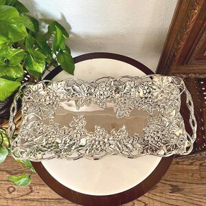 May include: A rectangular silver-toned metal serving tray featuring an intricate, raised relief border of grape clusters and vine leaves. The tray has a smooth, reflective central surface and integrated handles on both ends with a vine-like design.