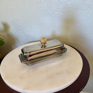 May include: A rectangular silver-toned metal butter dish with a polished finish and a tiered base. The lid features a small, textured gold-toned acorn-shaped handle centered on top, providing a decorative accent to the metallic container.