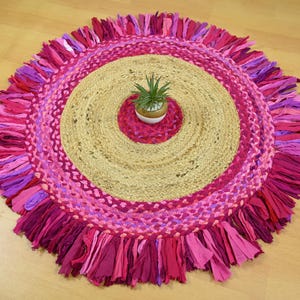 May include: Round rug with a jute center and concentric rings of pink and purple fabric. The outer edge features a fringe of pink fabric strips. A small potted plant sits in the center of the rug.