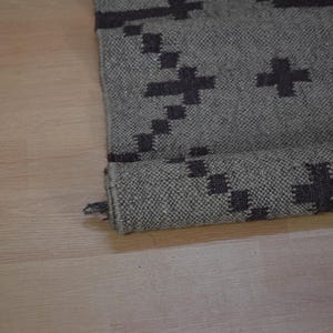 May include: Rolled-up gray rug with a dark gray geometric pattern. The rug is made of a woven material and has a rolled edge. The pattern consists of cross-like shapes.