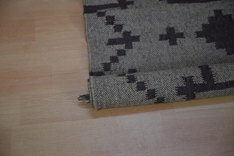 May include: Rolled-up gray rug with a dark gray geometric pattern. The rug is made of a woven material and has a rolled edge. The pattern consists of cross-like shapes.
