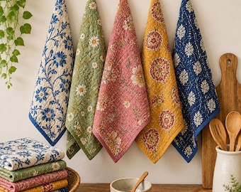 May include: Five quilted kitchen towels hanging in a row, featuring floral and geometric patterns in blue, green, pink, and yellow. A matching stack of folded towels sits in a woven basket, alongside a ceramic bowl and wooden utensils.
