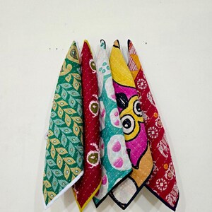 May include: A collection of six colorful, patterned fabric napkins hanging against a white wall. The napkins feature various designs, including leaf patterns, floral motifs, and abstract shapes in shades of green, red, yellow, and blue. Each napkin is folded into a triangle.