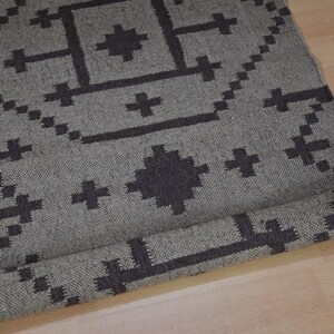 May include: A rolled-up gray rug with a geometric pattern in dark gray. The rug features cross and diamond shapes. The rug is made of woven material and is partially rolled up, revealing the underside.