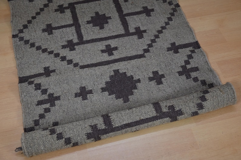 May include: A rolled-up gray rug with a geometric pattern in dark gray. The rug features cross and diamond shapes. The rug is made of woven material and is partially rolled up, revealing the underside.