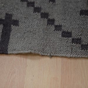 May include: Close-up of a gray rug with a repeating cross pattern in dark gray. The rug has a textured surface and a fringed edge. The rug is placed on a light brown wooden surface.