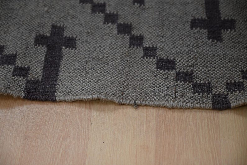 May include: Close-up of a gray rug with a repeating cross pattern in dark gray. The rug has a textured surface and a fringed edge. The rug is placed on a light brown wooden surface.