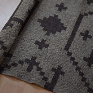 May include: A rolled-up, gray rug with a geometric pattern of dark gray crosses and lines. The rug is made of a woven material and is partially rolled, revealing the underside. The design is symmetrical and modern.