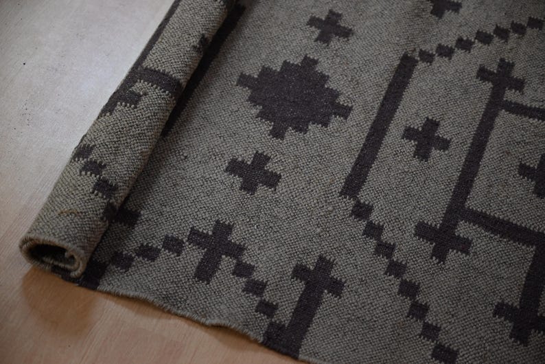 May include: A rolled-up, gray rug with a geometric pattern of dark gray crosses and lines. The rug is made of a woven material and is partially rolled, revealing the underside. The design is symmetrical and modern.