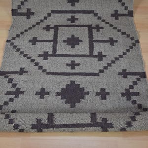 May include: A rolled-up gray rug with a geometric pattern in dark gray. The design features diamond and cross shapes. The rug is made of woven material and is displayed on a light wood surface.