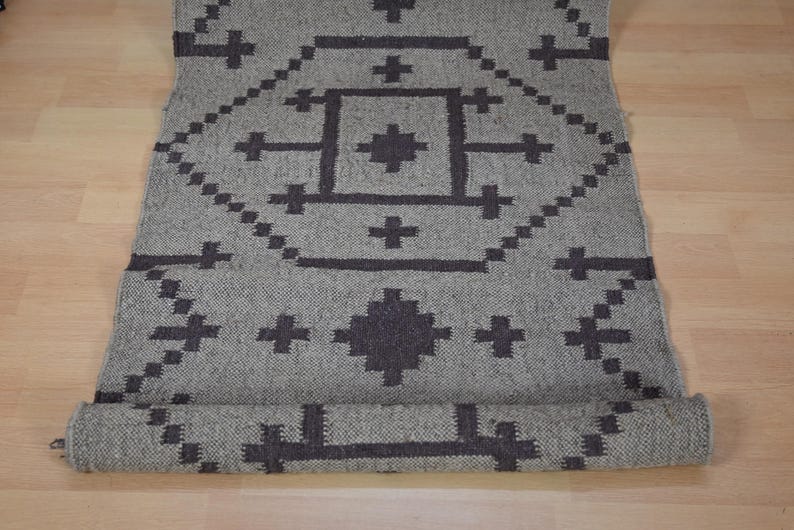May include: A rolled-up gray rug with a geometric pattern in dark gray. The design features diamond and cross shapes. The rug is made of woven material and is displayed on a light wood surface.