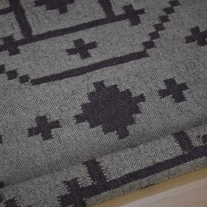 May include: A gray woven rug with a geometric pattern in dark gray. The design features cross-like shapes and rectangular lines. The rug is rolled up at the bottom, showing the texture of the fabric.