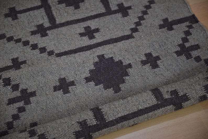 May include: A gray woven rug with a geometric pattern in dark gray. The design features cross-like shapes and rectangular lines. The rug is rolled up at the bottom, showing the texture of the fabric.