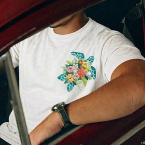 May include: White t-shirt featuring a teal turtle design with a floral arrangement. The turtle is embroidered with a variety of colorful flowers. The person is wearing a watch with a black band.